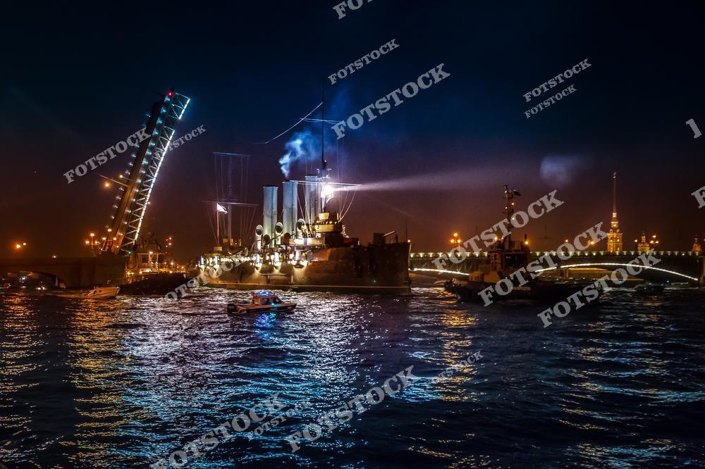 На фотографии изображён крейсер "Аврора", освещённый яркими ночными огнями. Виден разведённый мост, который подчёркивает динамичность ночной жизни Санкт-Петербурга. На заднем плане можно различить Петропавловскую крепость, освещённую тёплыми огнями. Крейсер "Аврора" – это не только символ города, но и музей, который хранит память о событиях революции.